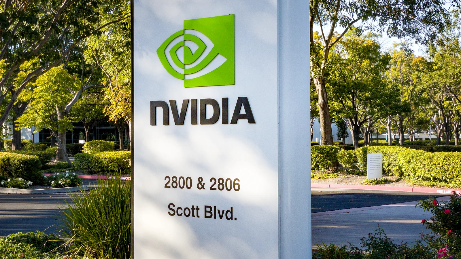 A tall white freestanding business sign displays a green eye-shaped logo, the word “NVIDIA,” and the address “2800 & 2806 Scott Blvd.” It stands in landscaped grounds with trees…